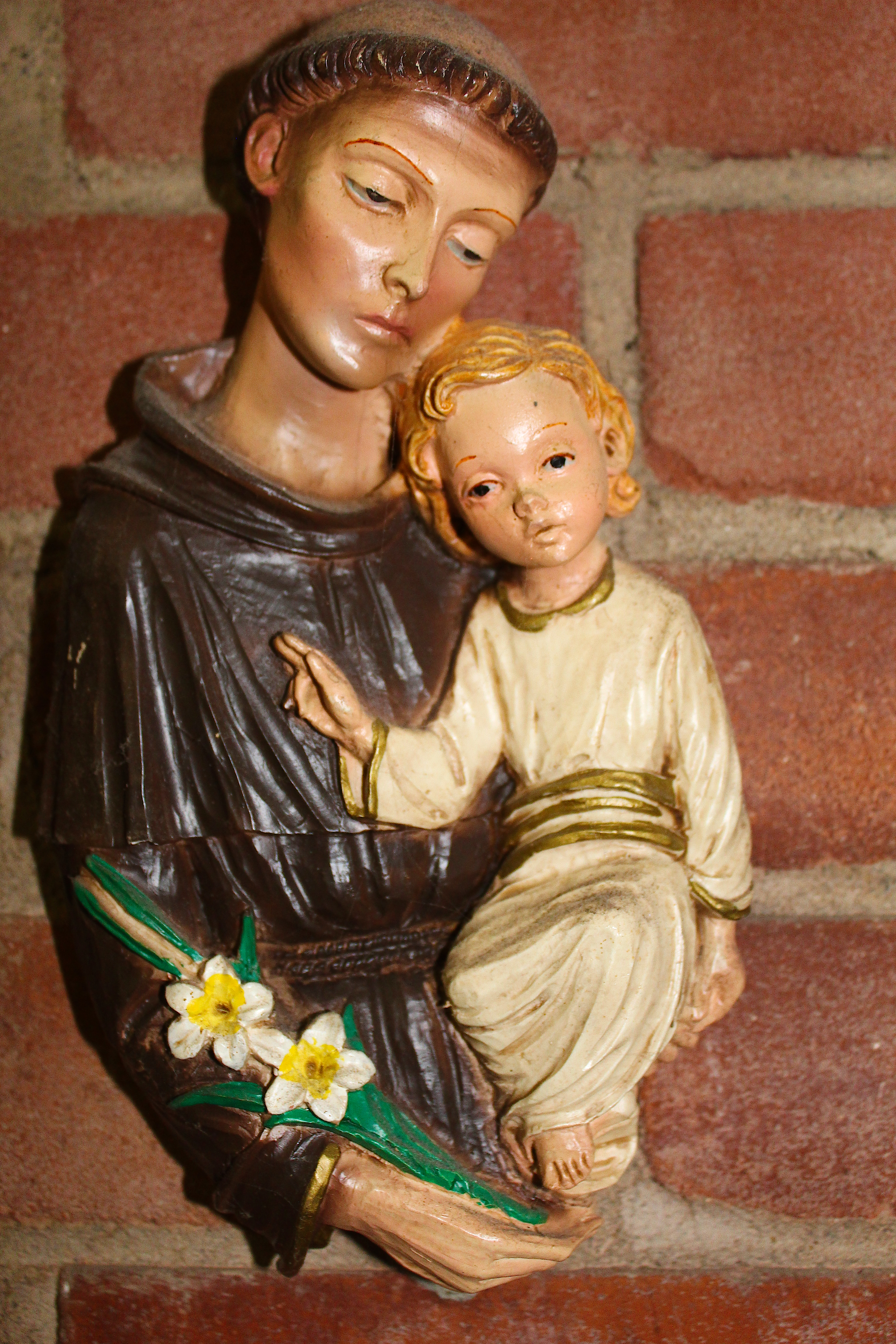 A small wall mounted hanging statur of St. Francis against a birck wall, holding daffodils and a baby Jesus