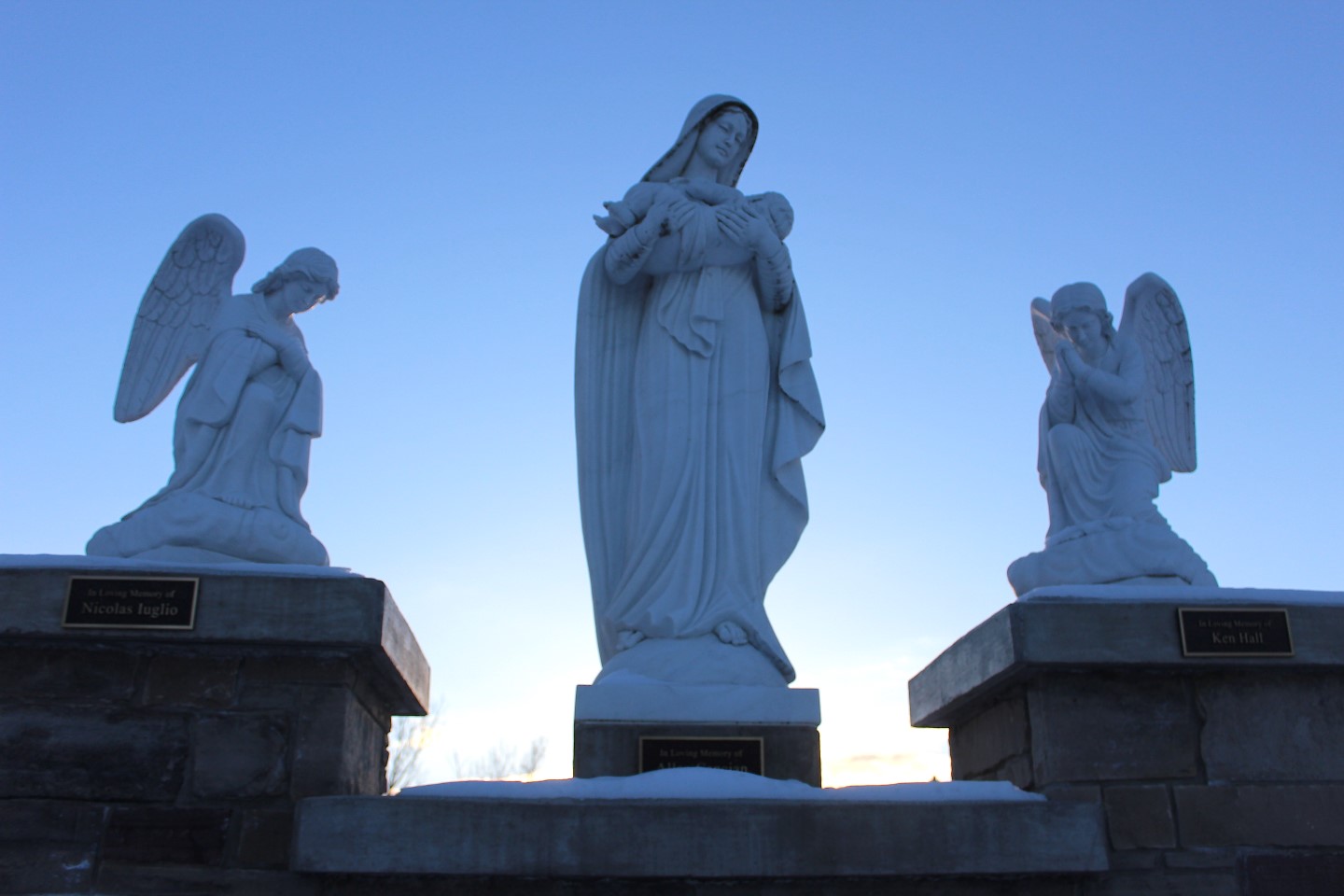 Cemetery monument with 3 large marble statues. 1 Angels on each sied, kneeling and looking into a Statue of the Virgin Mary holding the baby Jesus